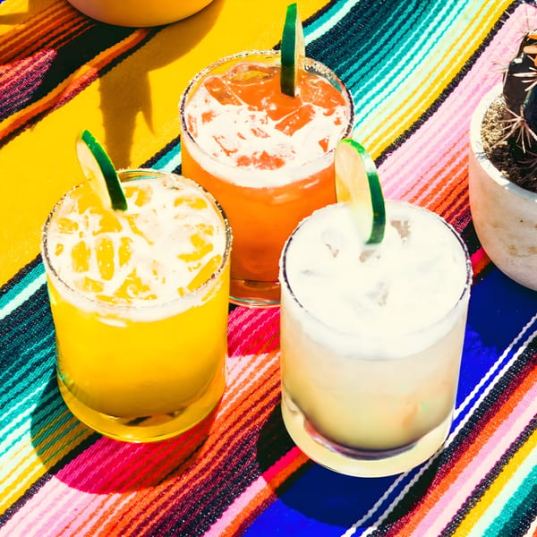 Three colorful margaritas with lime garnishes on a vibrant striped Mexican serape blanket.