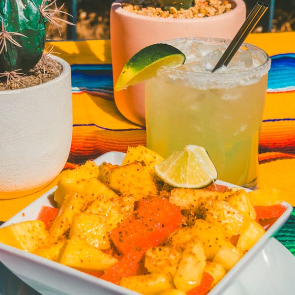 A vibrant Mexican fruit salad with chili powder next to a margarita on a colorful striped blanket.