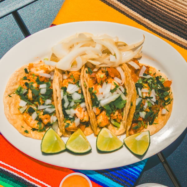 Four Tacos El Peladito tacos with chicken, onions, cilantro, and lime wedges on a white plate.