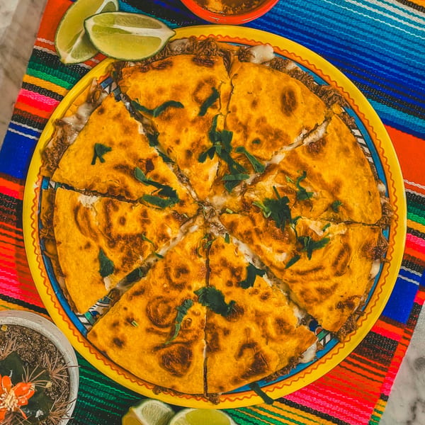 A large, sliced quesadilla with meat and cheese, garnished with cilantro and lime wedges on a colorful Mexican serape.