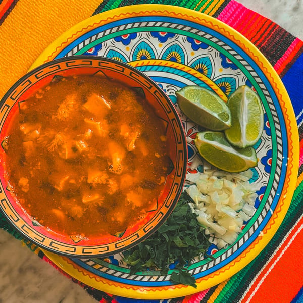 Menudo soup in a colorful bowl with limes, chopped onions, and cilantro on a vibrant plate.